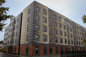 A large brick apartment complex at the corner of a street with a patterned mix of tan and black bricks. The complex has tall and narrow windows arranged in vertical columns across the complex.