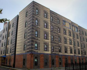 A large brick apartment complex at the corner of a street with a patterned mix of tan and black bricks. The complex has tall and narrow windows arranged in vertical columns across the complex.