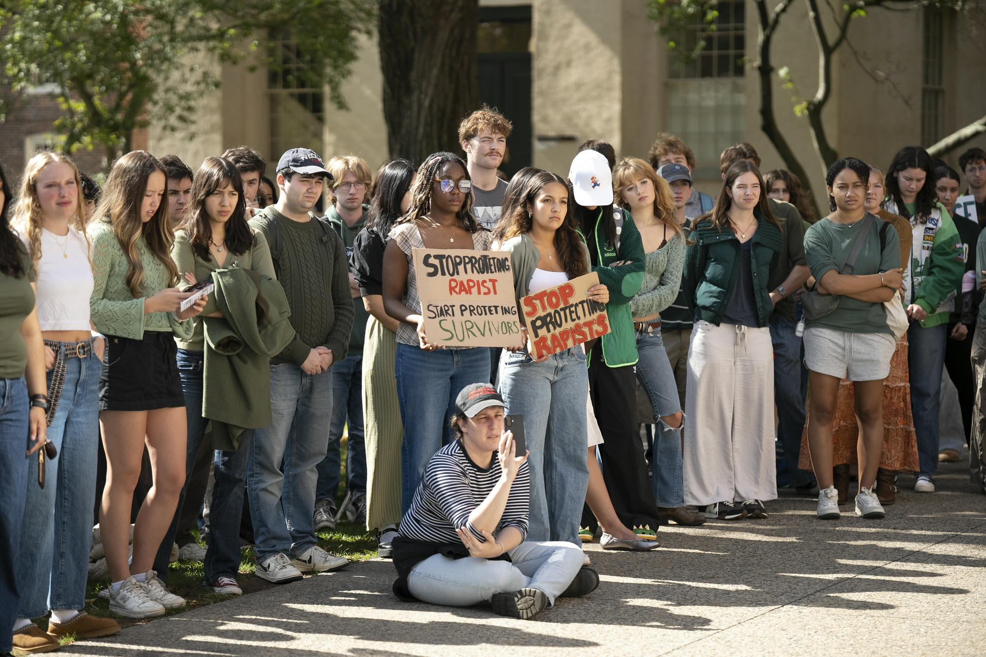 A photo of protestors standing on main green. Two protestors in the center of the photo are holding signs that read “Stop protecting rapists, start protecting survivors” and “Stop protecting rapists.” Someone is sitting in front of protestors recording on their phone.