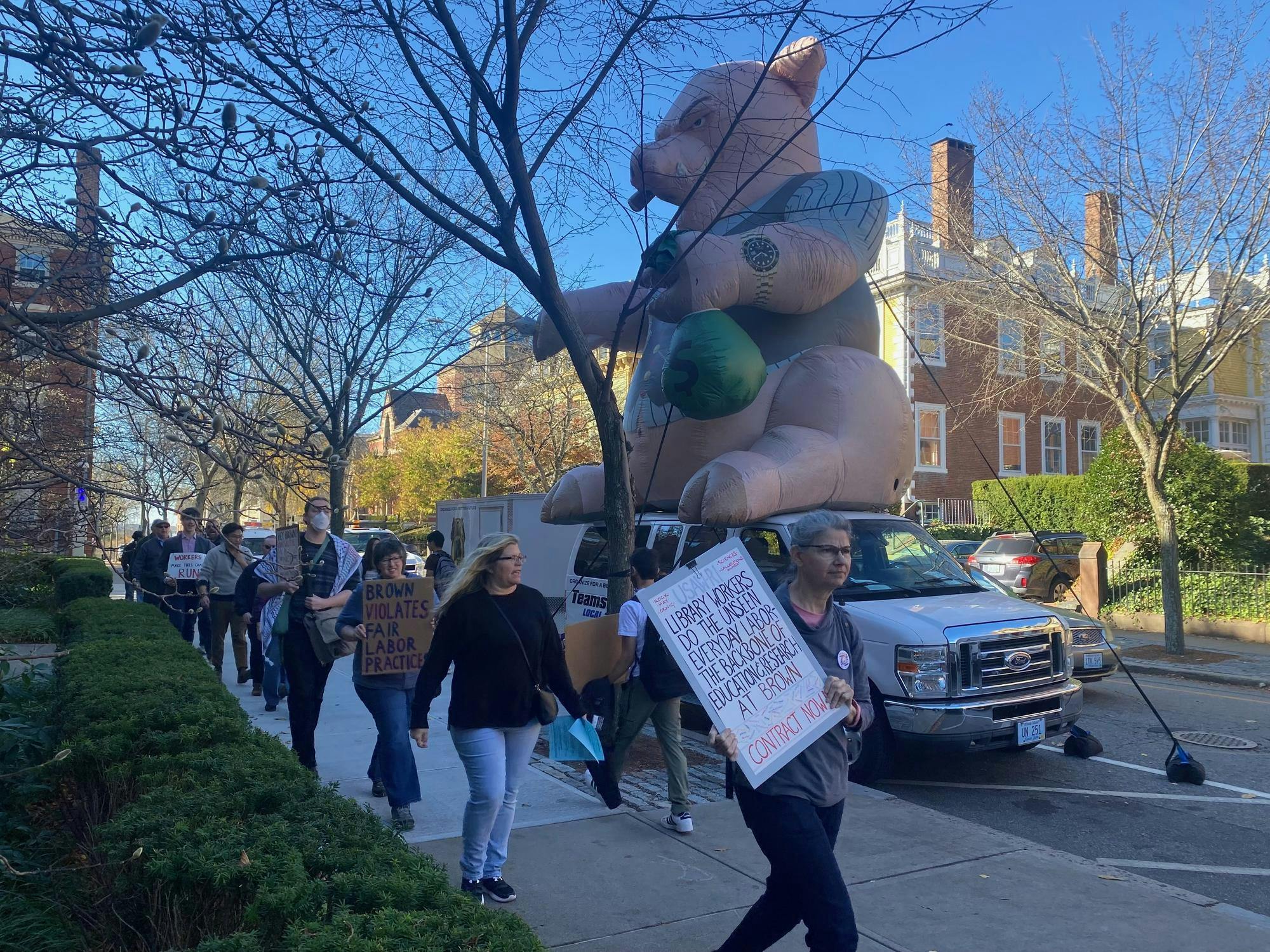 People holding signs reading "Brown violates fair labor practices" in front of Teamsters van that is carrying a large inflatable rat. This is taking place on Waterman Street near Faunce Arch. 