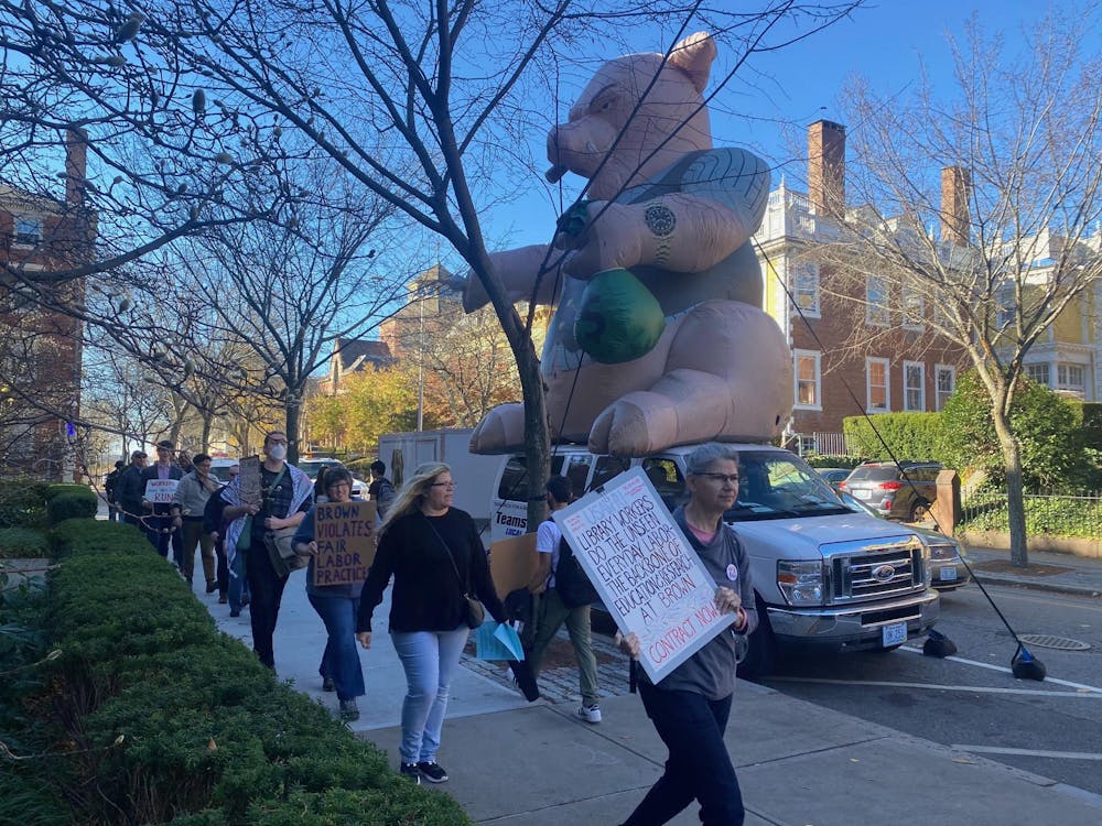 People holding signs reading "Brown violates fair labor practices" in front of Teamsters van that is carrying a large inflatable rat. This is taking place on Waterman Street near Faunce Arch. 