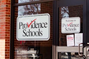 The window of the Providence Schools building. There are 2 signs that say Providence Schools in black text with a red checkmark for the "v" in "Providence."