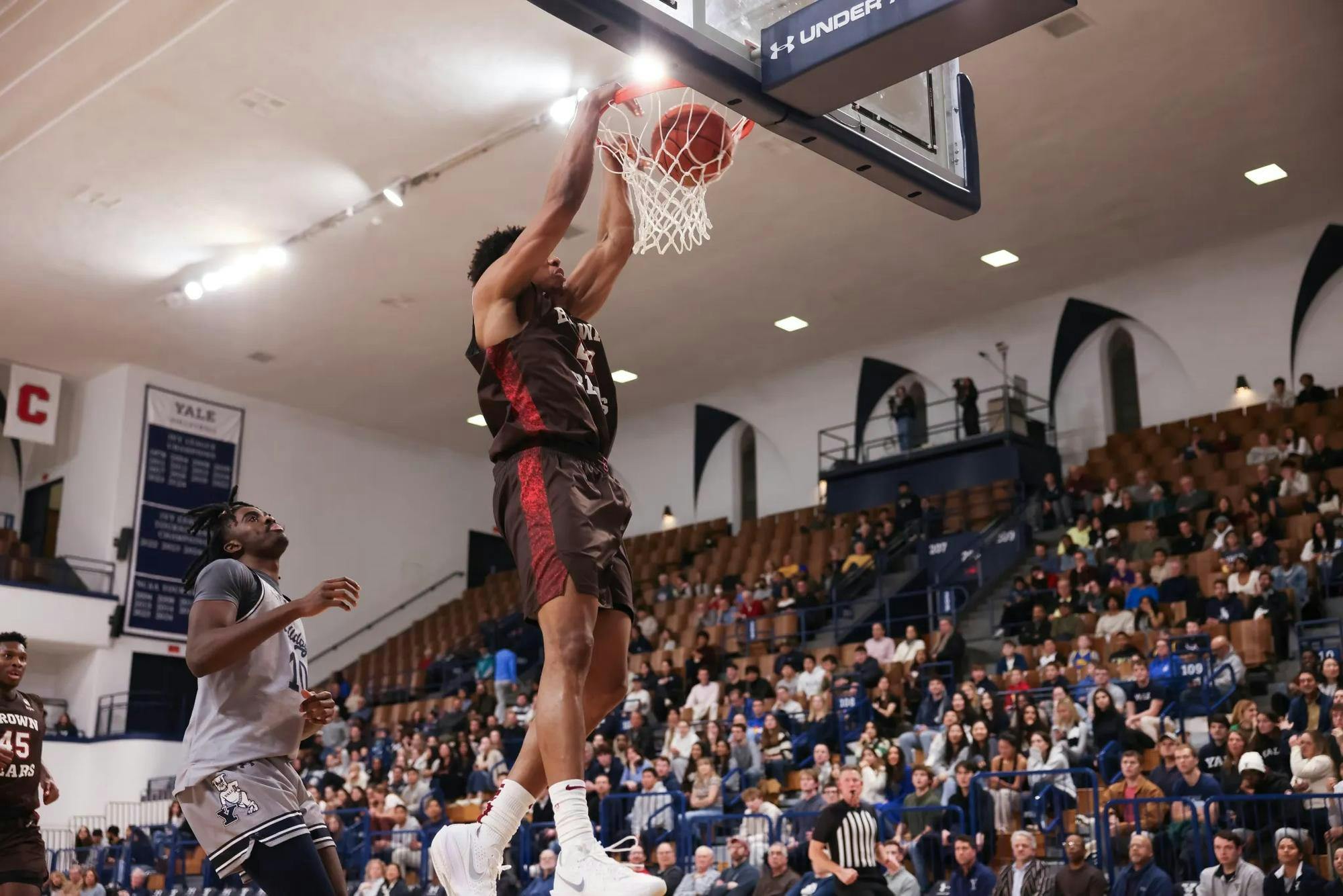 Brown team player dunking mid-air with a Yale team player behind him and a crowd of fans in the background.