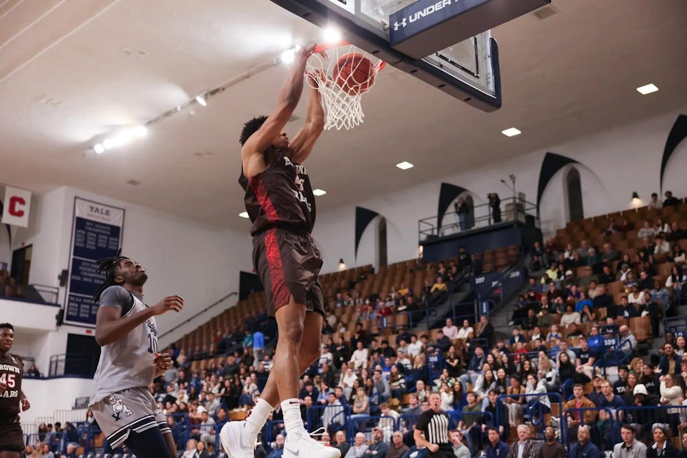 Brown team player dunking mid-air with a Yale team player behind him and a crowd of fans in the background.
