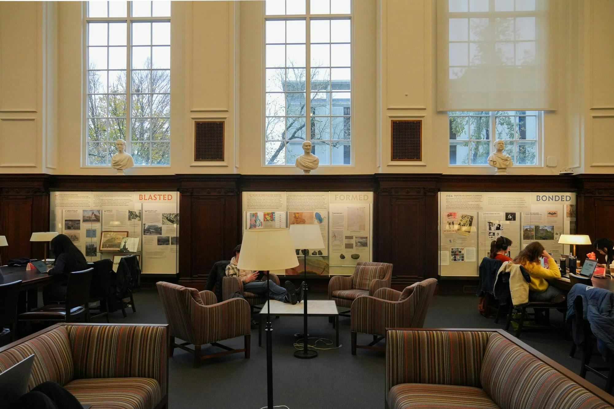 Photo of the "Shared Magma: An American and Swedish Collaboration on Sisterhood and Sister Ore” exhibit at the John Hay Library viewed from across the room, with students working at tables and chairs in front of and to the side of three shadowbox galleries displaying elements of the exhibit.