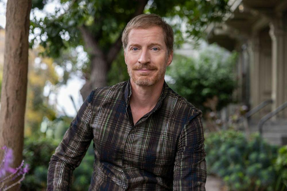 Photo of Andrew Sean Greer in a plaid shirt sitting in front of green foliage.