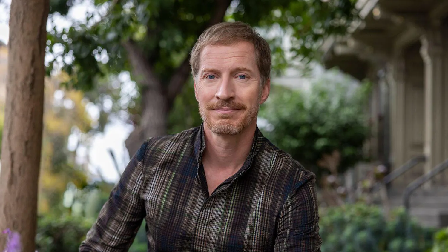 Photo of Andrew Sean Greer in a plaid shirt sitting in front of green foliage.