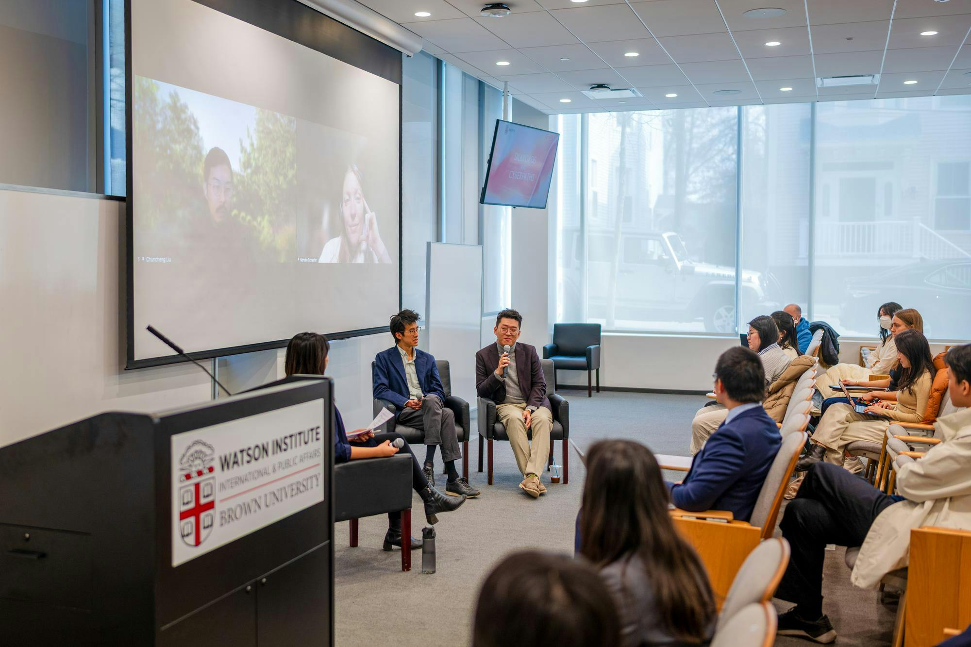 A picture of the audience and speakers at the Brown China Summit. There are two speakers on Zoom on a projector screen in the back.