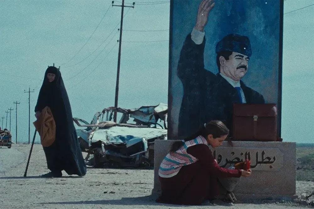 Photo of a girl looking at something she's holding while she kneels in front of a large painting of a man gesturing with his hand. She is next to a street, and a woman in the background looks towards her.