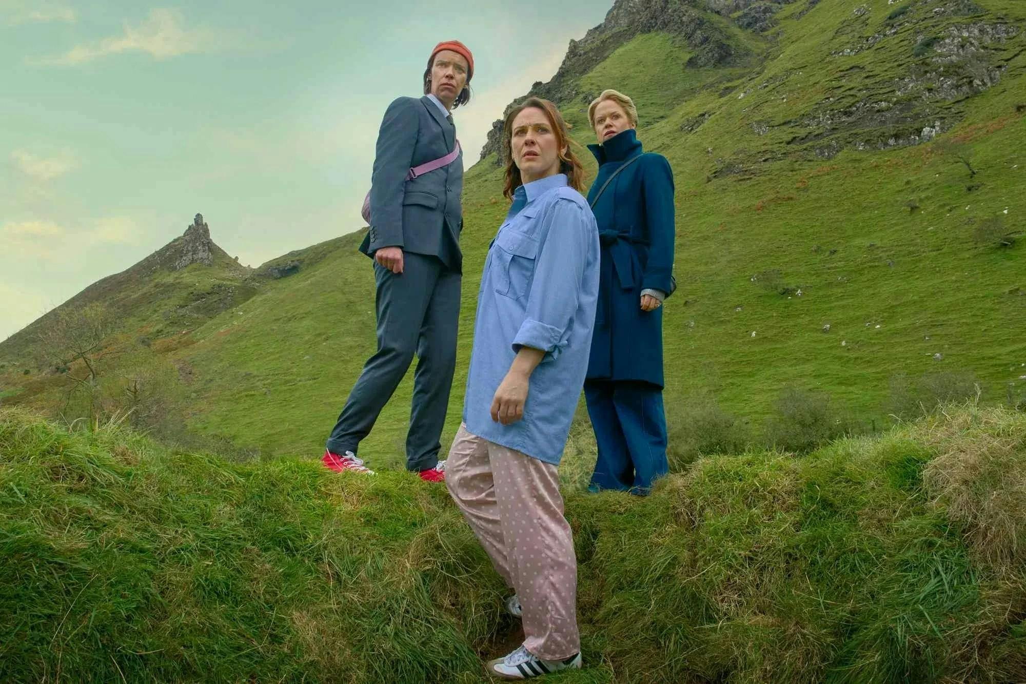 Three women standing on grass are pictured against a sprawling green mountainscape.


