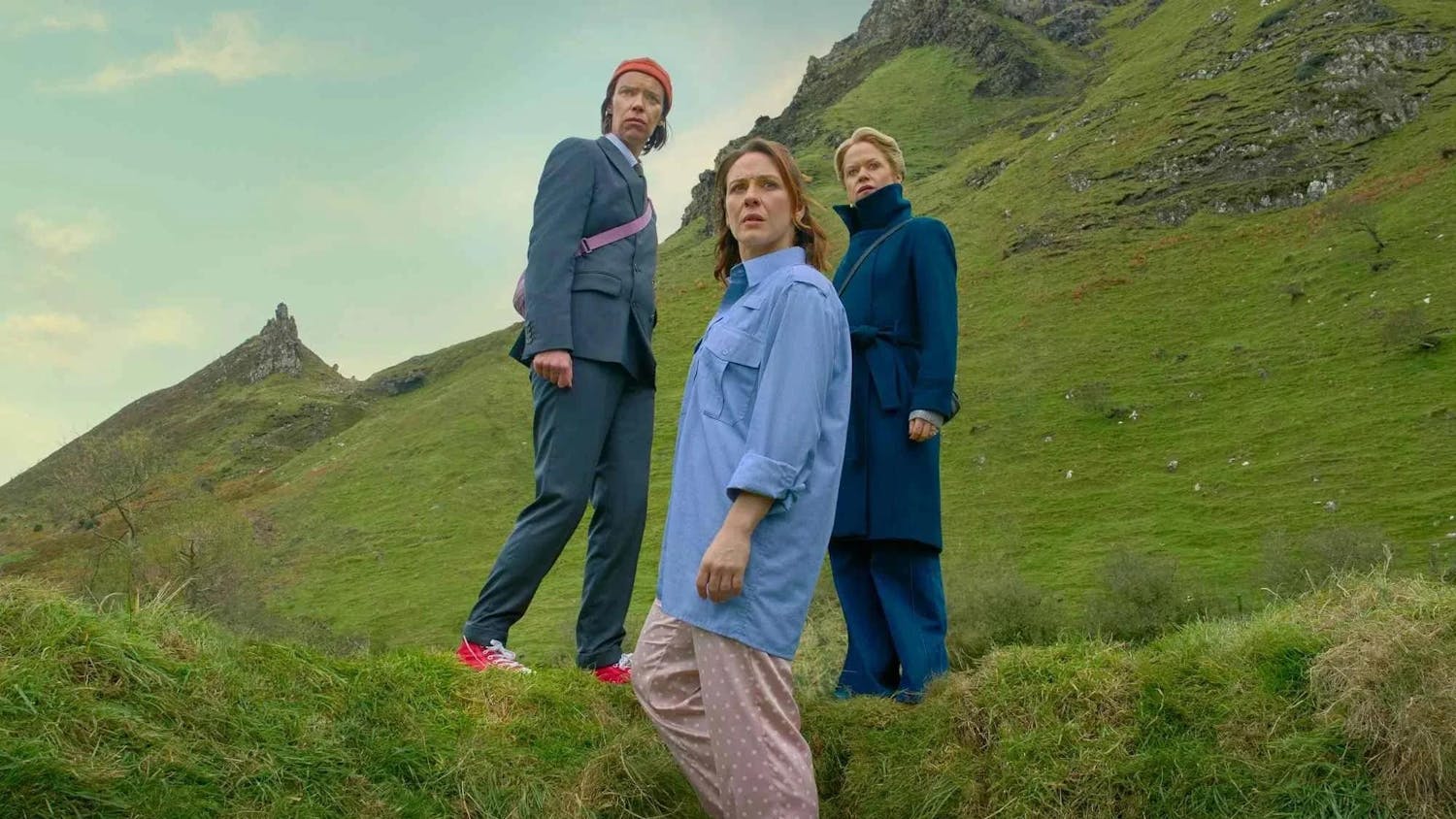 Three women standing on grass are pictured against a sprawling green mountainscape.