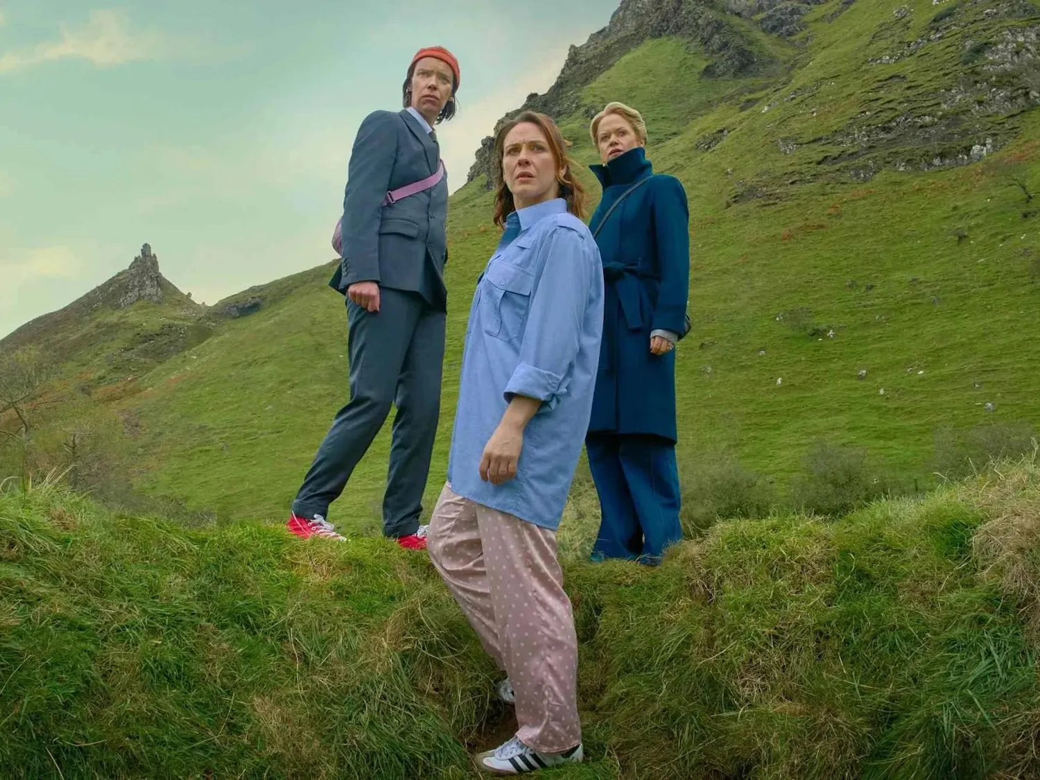 Three women standing on grass are pictured against a sprawling green mountainscape.