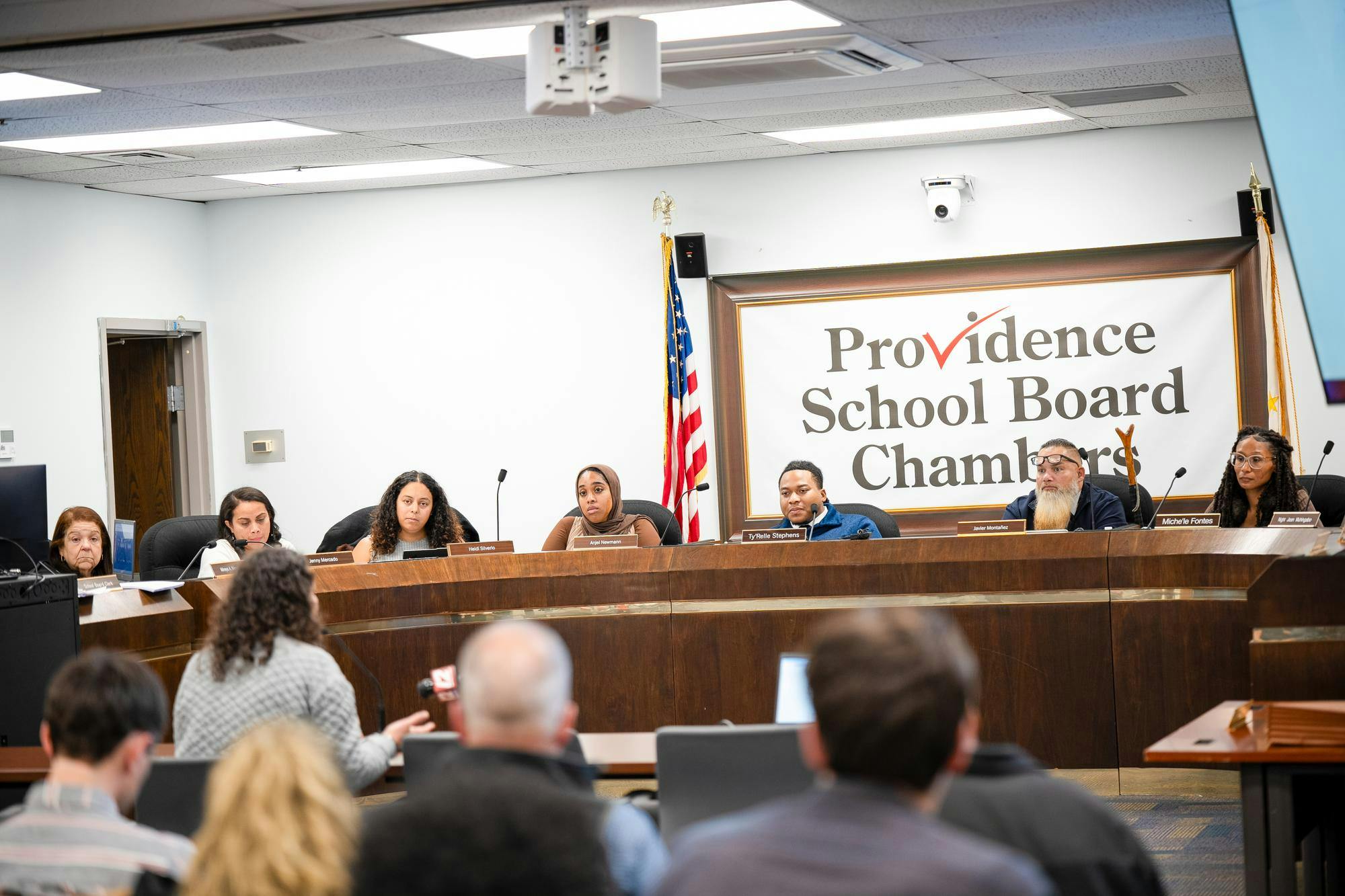 A photo of the Providence School Board sitting around a semi-circle wooden table facing a crowd of people. They watch one woman speak to them. 