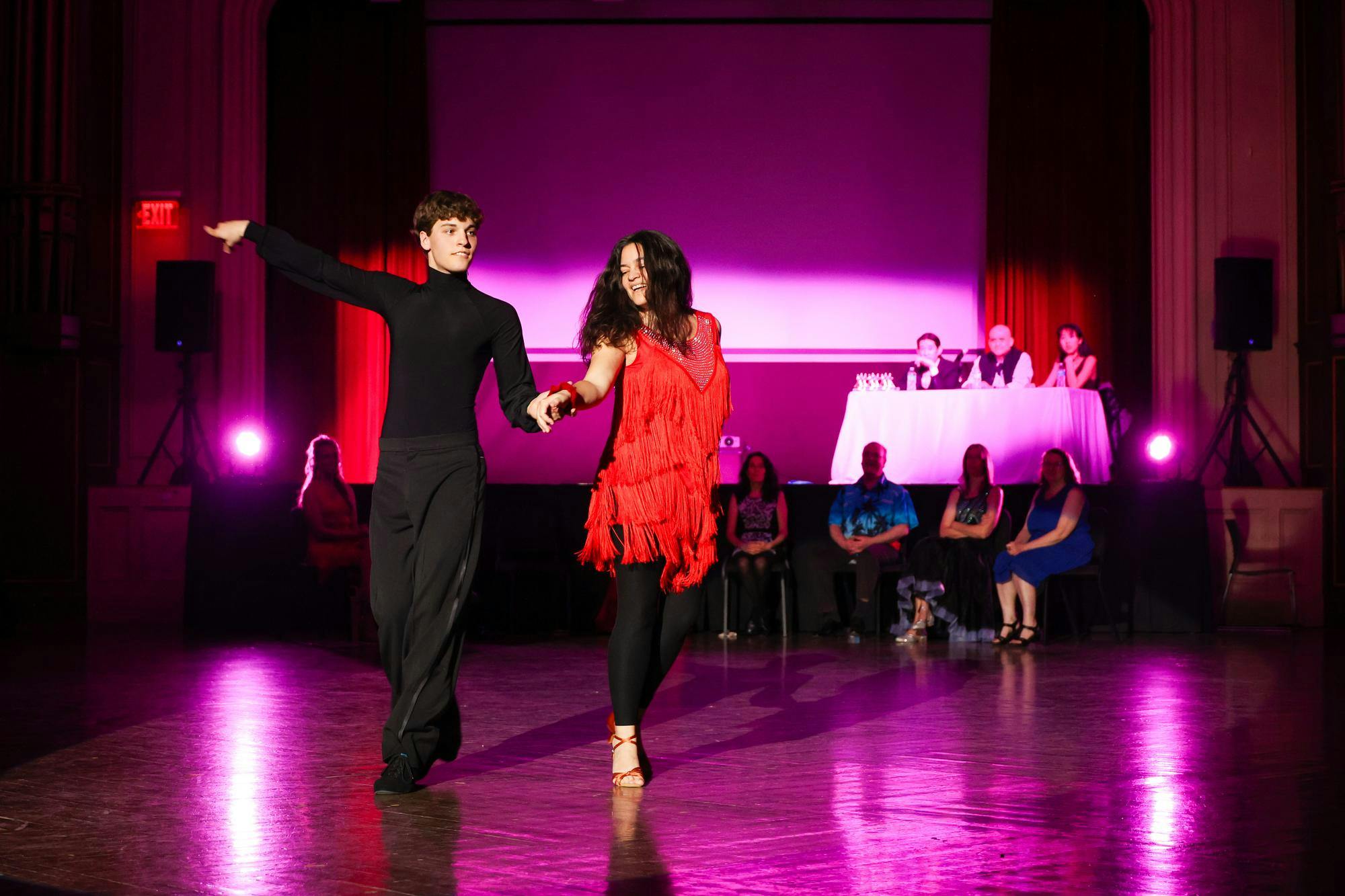 A man in all black dances with a woman in a red dress under pink light.