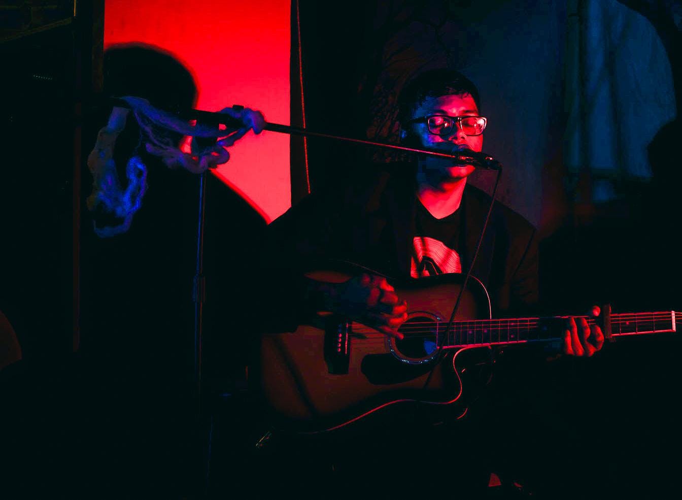 Photo of someone playing the guitar while singing with their eyes closed into a microphone in a dark room lit in pink and blue.
