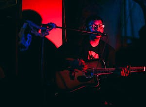 Photo of someone playing the guitar while singing with their eyes closed into a microphone in a dark room lit in pink and blue.