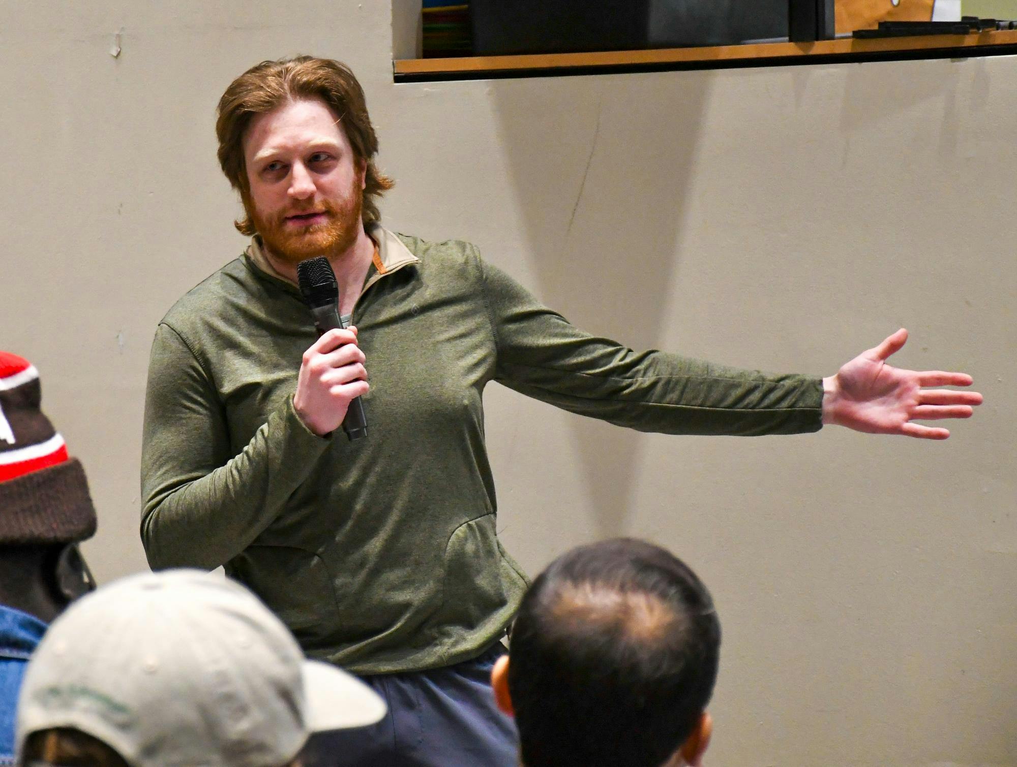A white man in a green long sleeved t-shirt with red hair and a beard holds a microphone with his mouth slightly opened. 