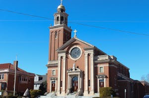 Photo of a brick building with a bell tower and crosses on the roof and on top of the belltower with a circular window above a statue and a set of wooden double doors with white stairs leading up to the doors.