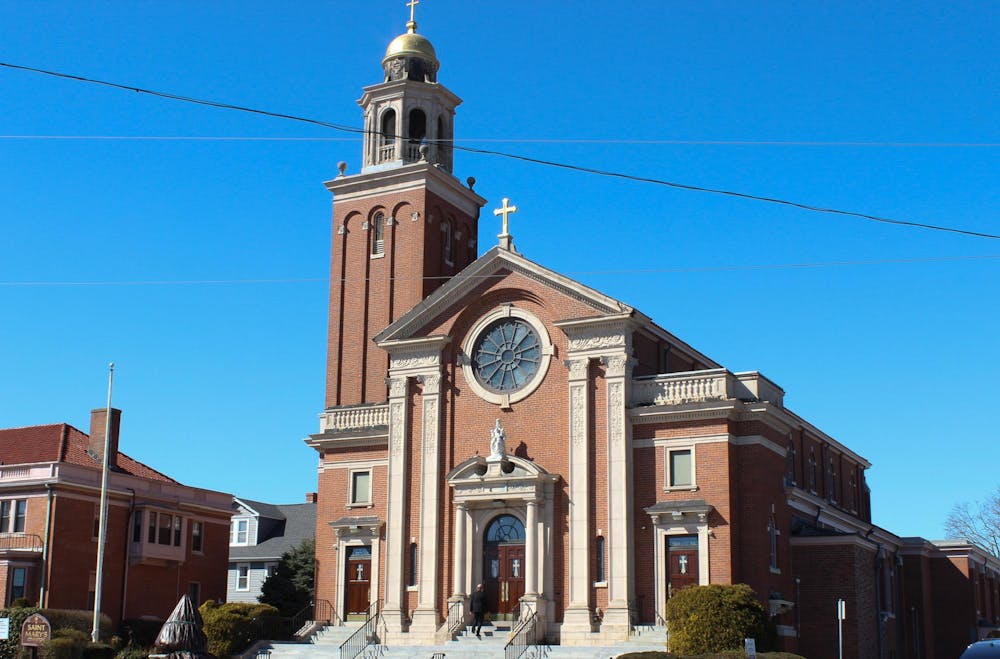 Photo of a brick building with a bell tower and crosses on the roof and on top of the belltower with a circular window above a statue and a set of wooden double doors with white stairs leading up to the doors.