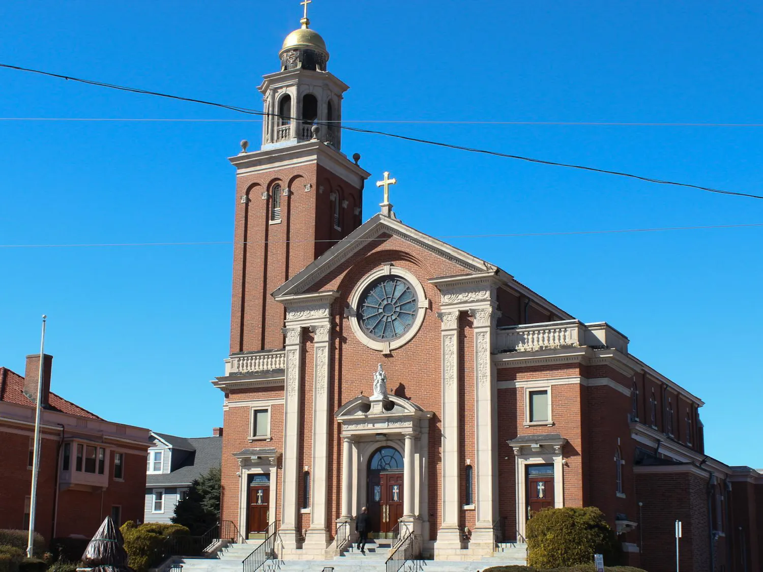 Photo of a brick building with a bell tower and crosses on the roof and on top of the belltower with a circular window above a statue and a set of wooden double doors with white stairs leading up to the doors.