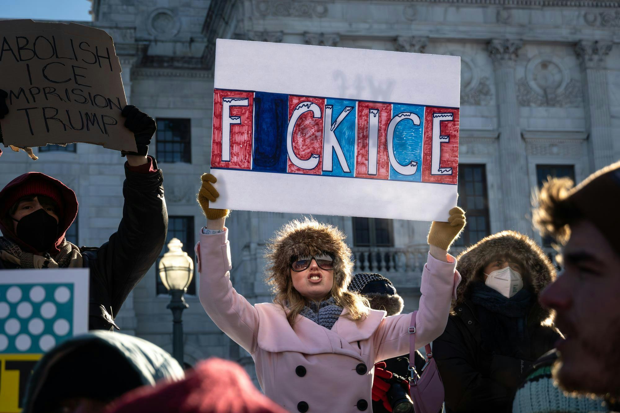 A woman in a pink coat, sunglasses, and a furry hat holds a sign that reads “Fuck Ice.”