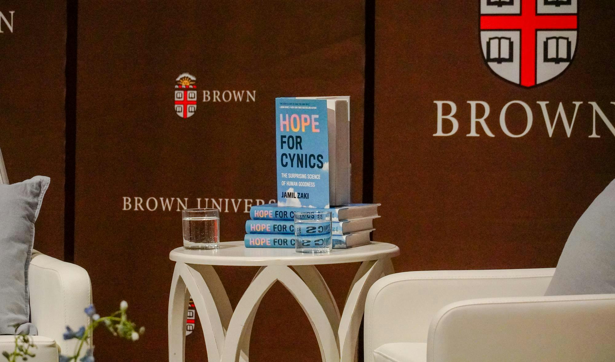 A photo of a table between two white chairs. On the table are copies of a book called "Hope for Cynics" with a blue cover and two glasses of water. 