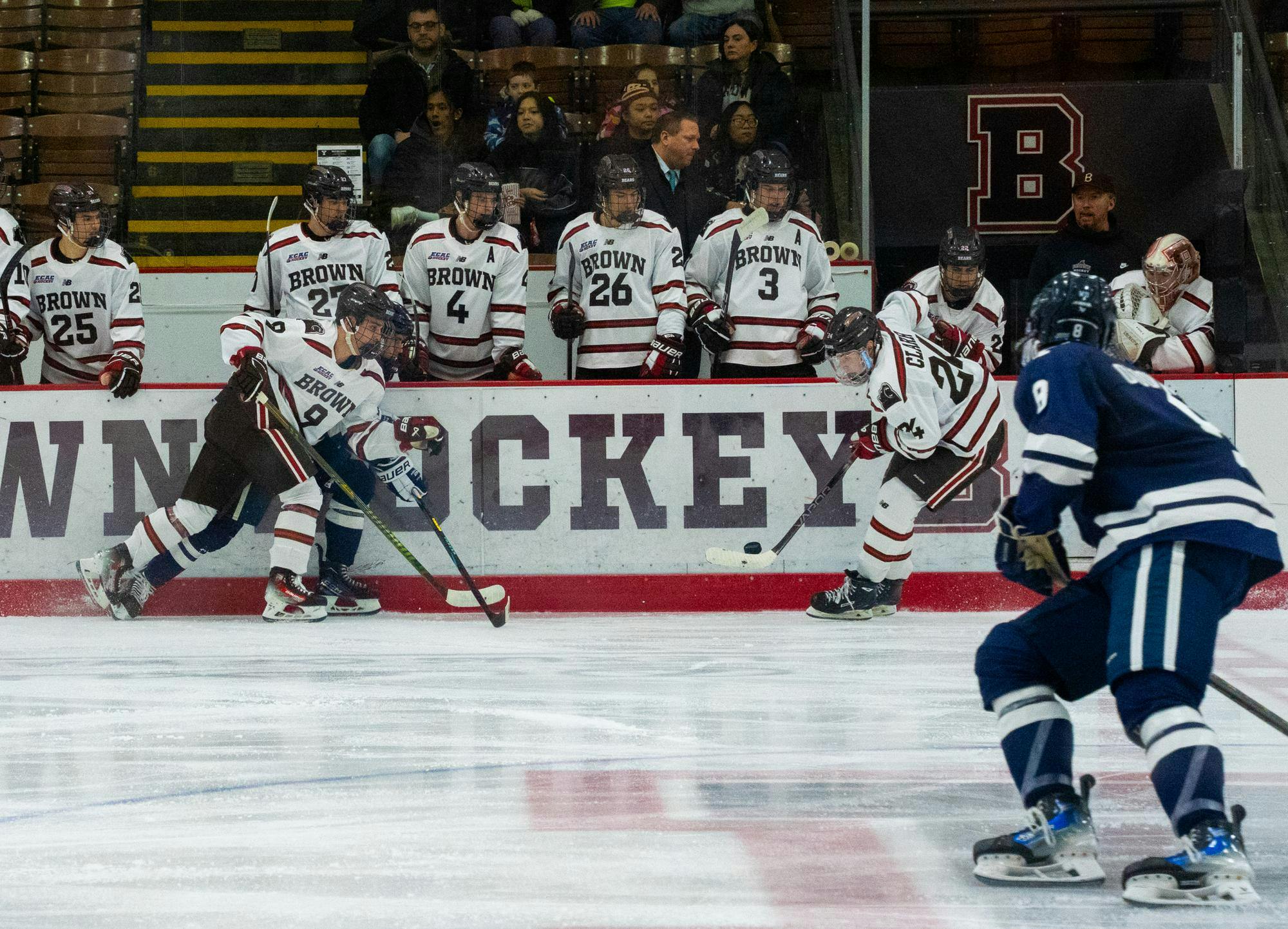 On the Meehan Auditorium ice, one Brunonian juggles the puck on his stick as two other teammates flood in to support him. In the foreground, a Yale Bulldog rushes into the play. The Bears stand just behind the wall, watching the play attentively.