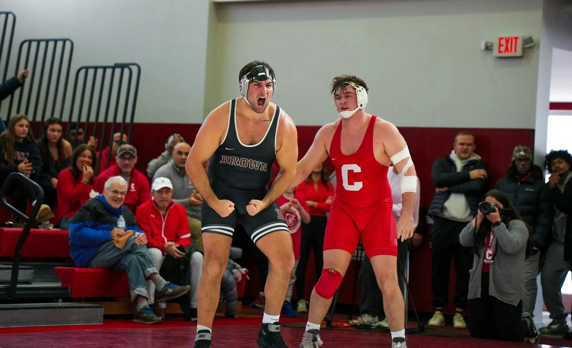 Brown wrestler standing besides Cornell wrestler with a crowd of fans in the background.