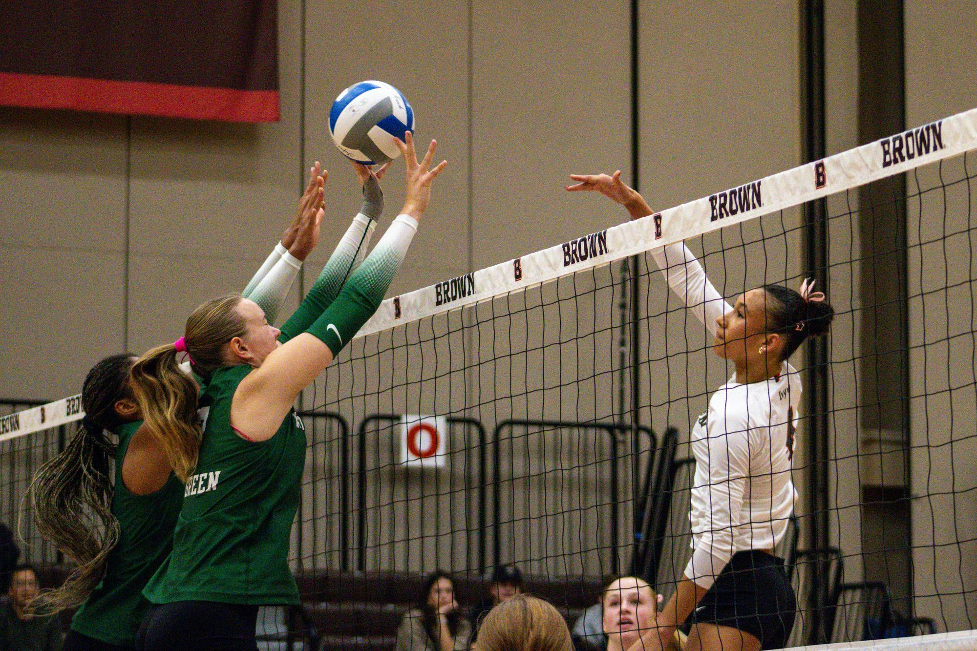 Photo of Brown women's volleyball's matchup against Dartmouth, as two Dartmouth players in green uniforms reach to block a ball which has just been sent over the net by a Brown volleyball player. 