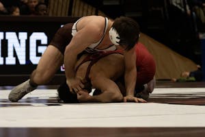 Photo of Brown wrestler in a singlet that is white on the top and brown on the legs and a big brown “B” on the chest, and a white helmet, curled over a Harvard wrestler in a red singlet and red helmet curled up on the ground.

