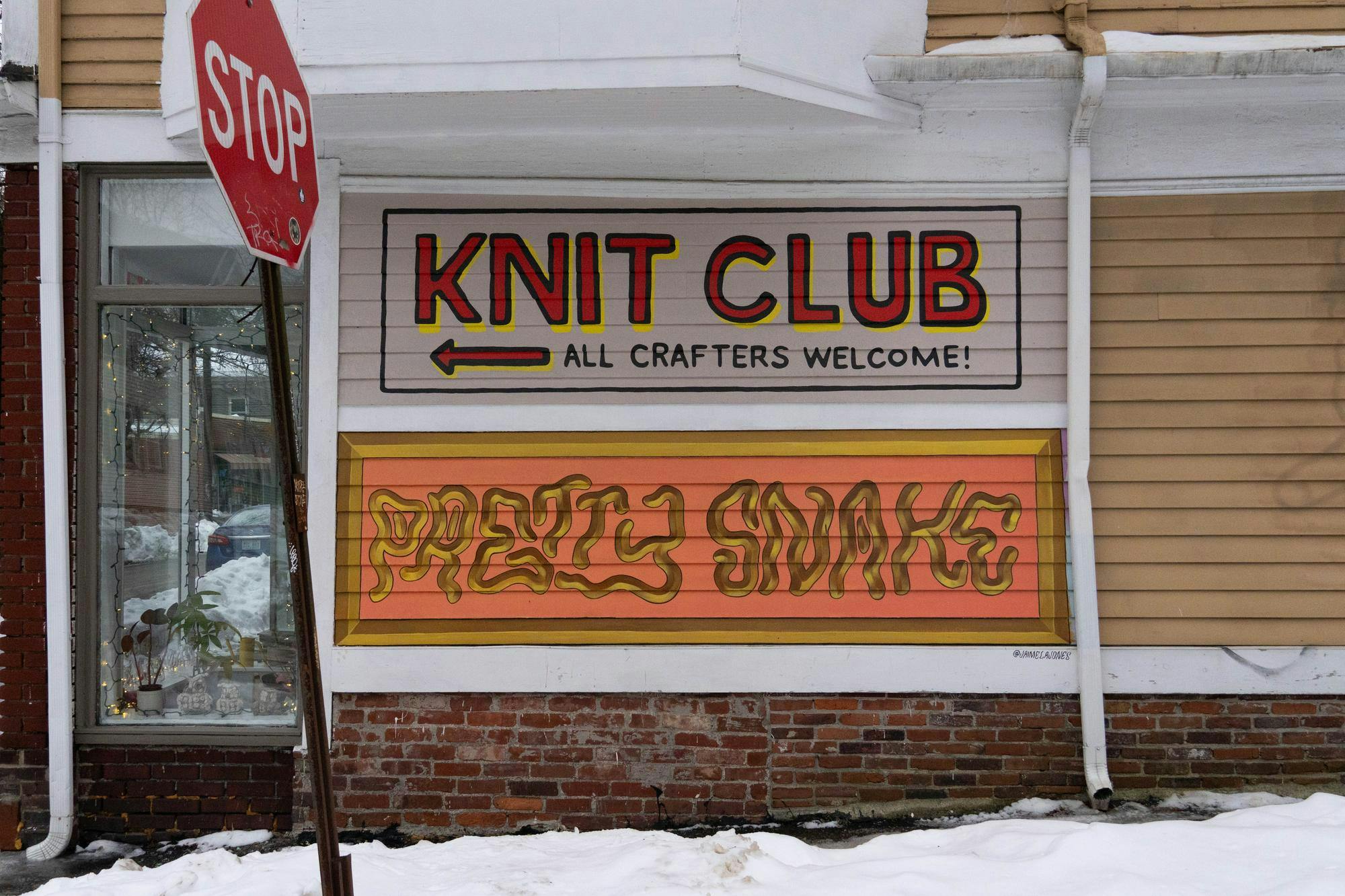 Wall text saying “Knit Club (left arrow) All Crafters Welcome!” and “Pretty Snake” with a stop sign on the front left. 

