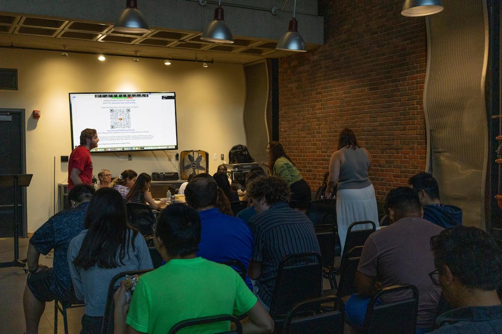 The photo depicts a meeting in progress, with a few rows of people with their backs to the camera. The front of the room holds a TV with a QR code displayed. All but two members are seated, with the standing two conversing from across a table in front of the rows of seats.