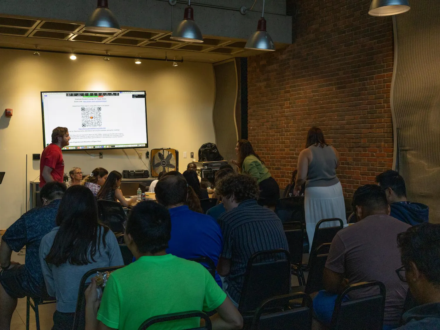 The photo depicts a meeting in progress, with a few rows of people with their backs to the camera. The front of the room holds a TV with a QR code displayed. All but two members are seated, with the standing two conversing from across a table in front of the rows of seats.