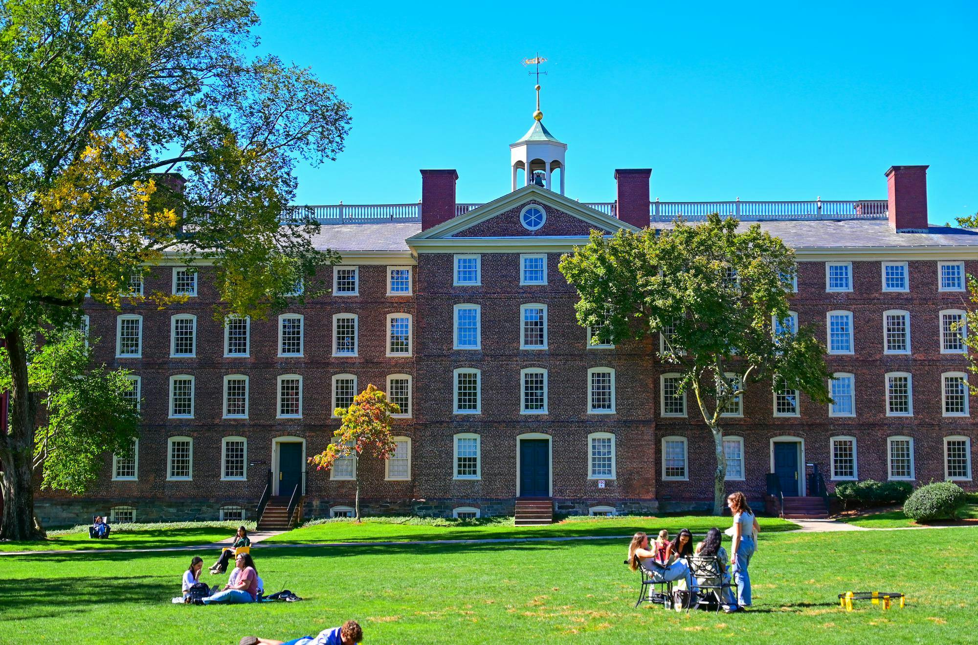 A photo of Brown University’s University Hall, a large brick building, with students in front of the building on the Main Green and a clear blue sky behind. 