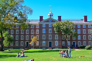 A photo of Brown University’s University Hall, a large brick building, with students in front of the building on the Main Green and a clear blue sky behind. 