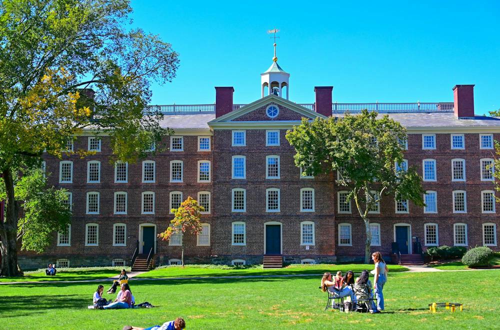 A photo of Brown University’s University Hall, a large brick building, with students in front of the building on the Main Green and a clear blue sky behind. 