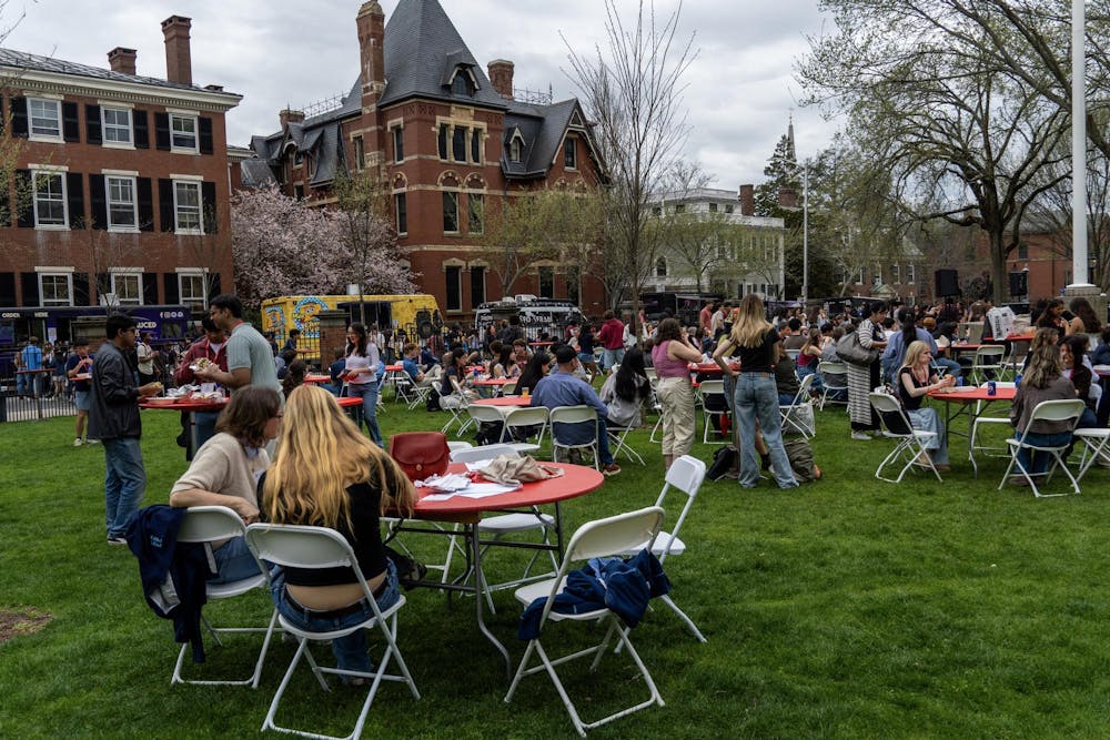A photo showing people during ADOCH sitting at chairs and tables on the Main Green with food trucks parked on the street in the background. 