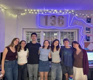 Seven beautiful individuals stand in front of a large sign that reads "136" illuminated by a neon blue glow. 