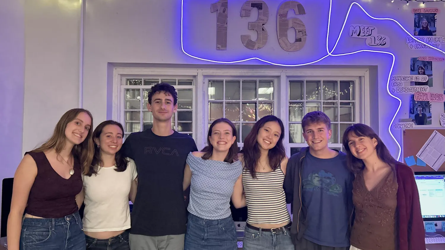 Seven beautiful individuals stand in front of a large sign that reads "136" illuminated by a neon blue glow.