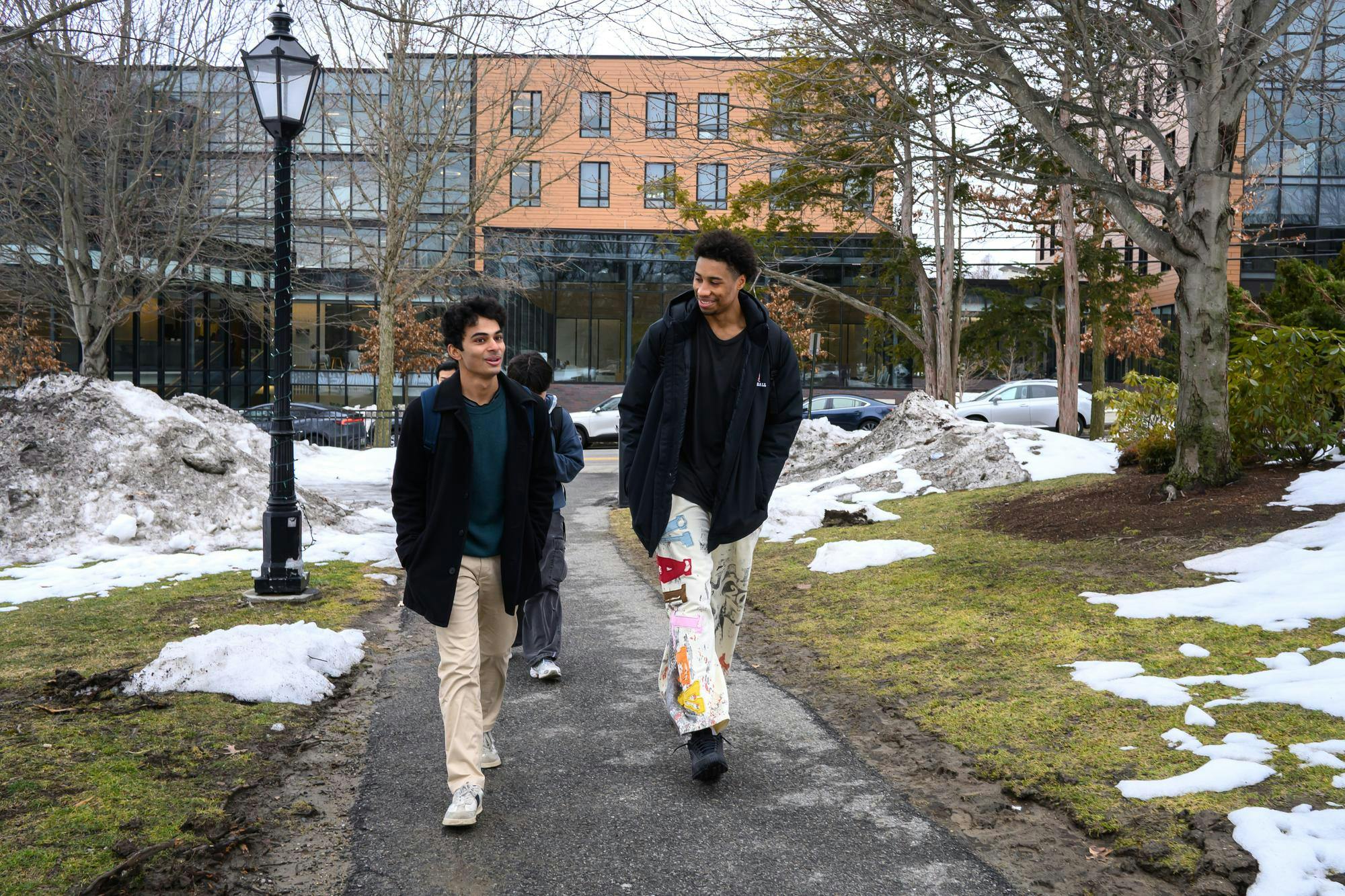 Lewis talks to another man as they walk through a park. There is snow and green grass behind them.