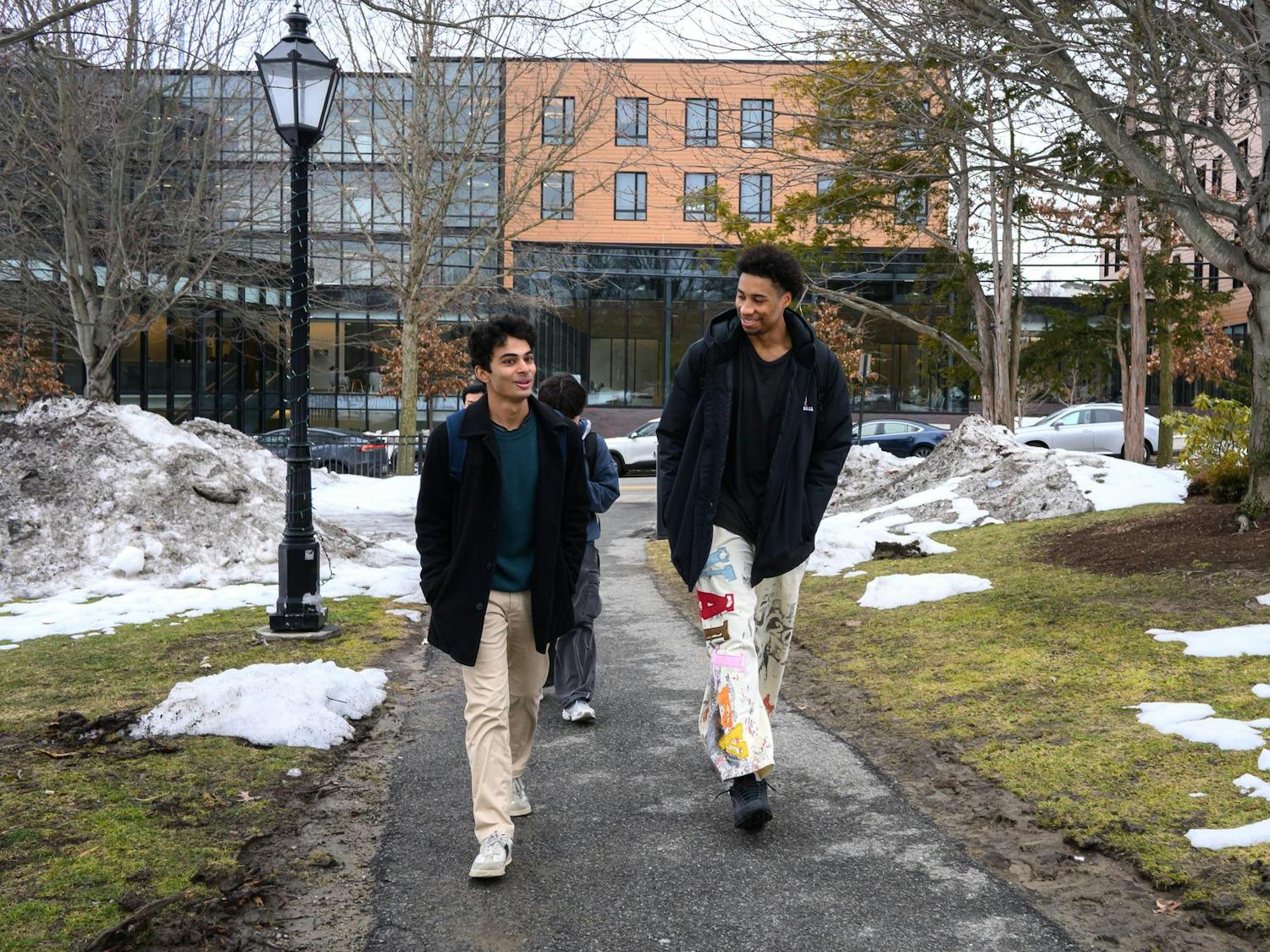 Lewis talks to another man as they walk through a park. There is snow and green grass behind them.