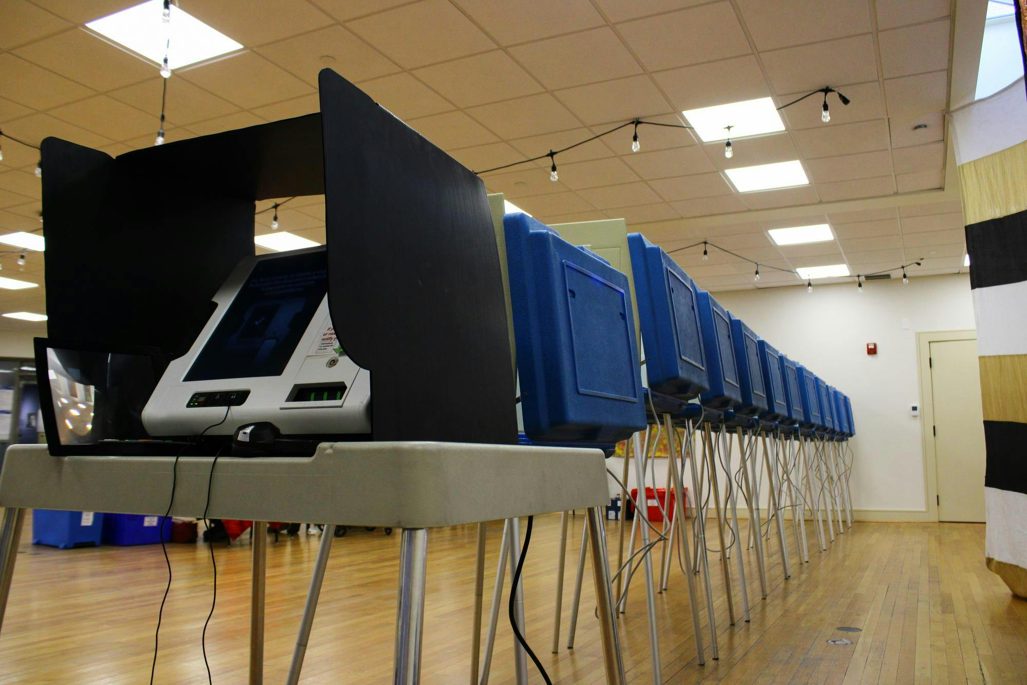 Photo of a voting machine on a white school desk with metal legs covered by a white privacy divider. Behind the desk, the backs of blue dividers are seen with wires dangling below.