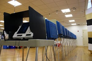 Photo of a voting machine on a white school desk with metal legs covered by a white privacy divider. Behind the desk, the backs of blue dividers are seen with wires dangling below.