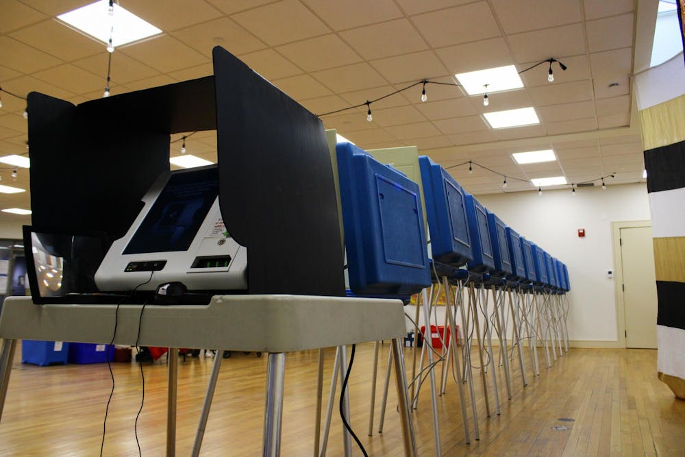 Photo of a voting machine on a white school desk with metal legs covered by a white privacy divider. Behind the desk, the backs of blue dividers are seen with wires dangling below.