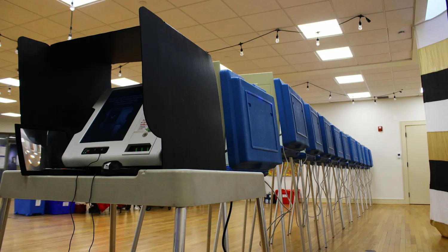 Photo of a voting machine on a white school desk with metal legs covered by a white privacy divider. Behind the desk, the backs of blue dividers are seen with wires dangling below.