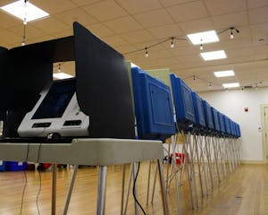 Photo of a voting machine on a white school desk with metal legs covered by a white privacy divider. Behind the desk, the backs of blue dividers are seen with wires dangling below.
