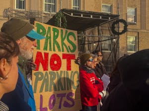 Around 6 demonstrators stand at a protest against the City of Pawtucket's plans to sell a portion of Morley Field, holding a multi-colored sign that reads "Parks not parking lots."