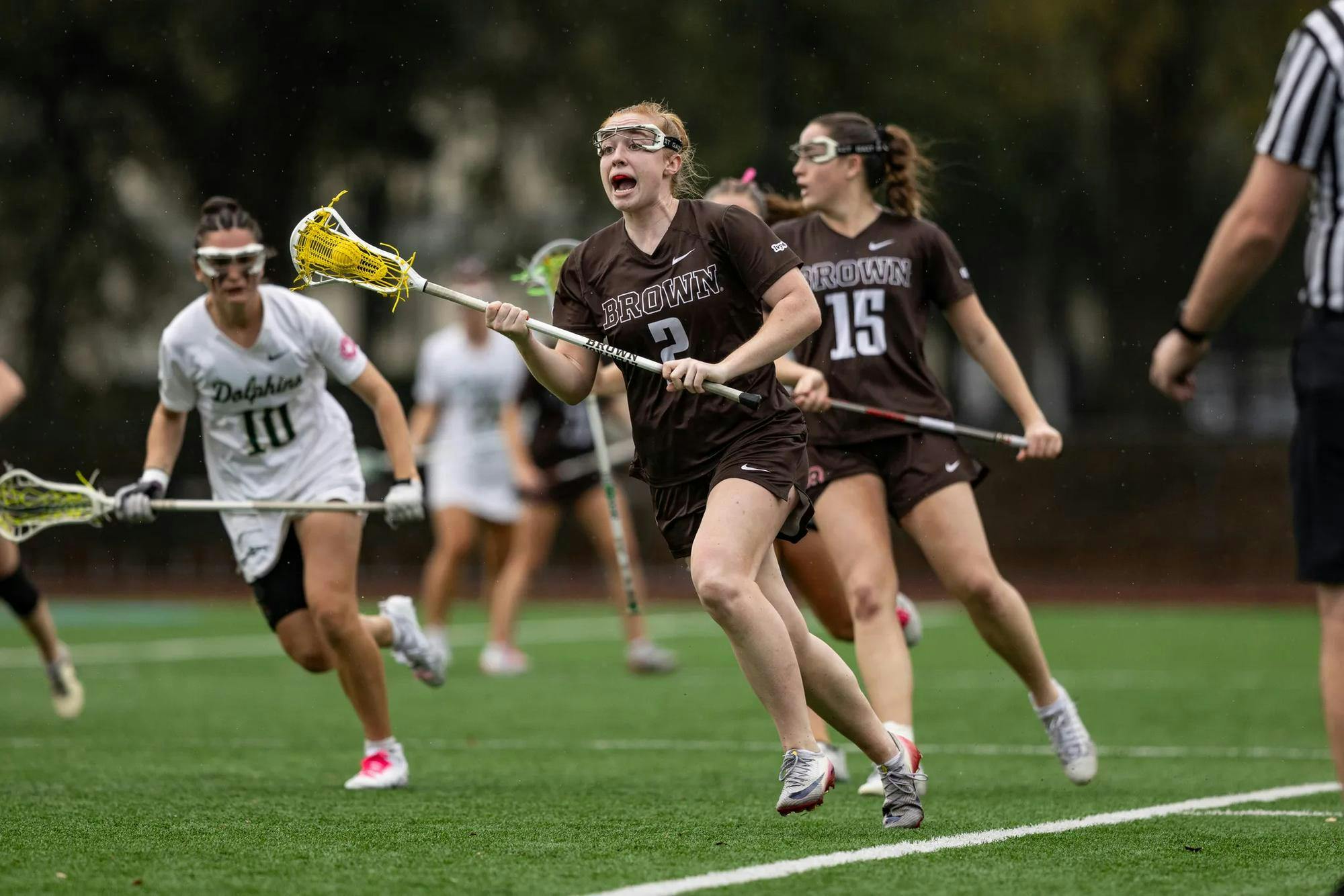 Photo of a female lacrosse player from Brown yelling while in possession of the ball with a Jacksonville player in white chasing behind.