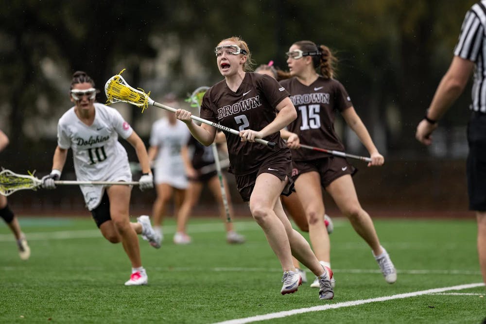 Photo of a female lacrosse player from Brown yelling while in possession of the ball with a Jacksonville player in white chasing behind.