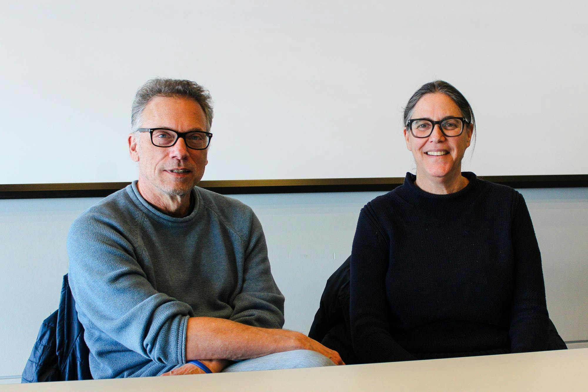 Visiting lecturers Jennifer Levin and John Eisendrath, wearing matching black-rimmed glasses and black and gray sweaters, smile against a white background. 
